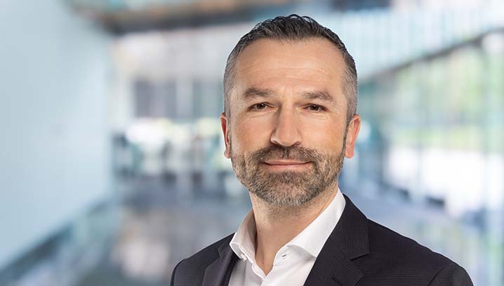 Portrait von Ado Bektas in schwarzem Sakko und weißem Hemd. Hintergrund unscharf, zeigt modernes Bürogebäude.